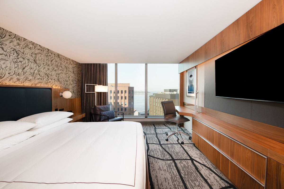 A modern hotel room with a king-size bed, desk, chair, and city view